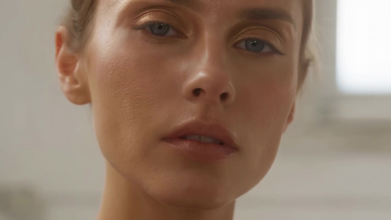 Tight portrait shows a woman looking toward camera, briefly closing and reopening her eyes as her head makes small natural movements, with warm makeup and soft indoor daylight.