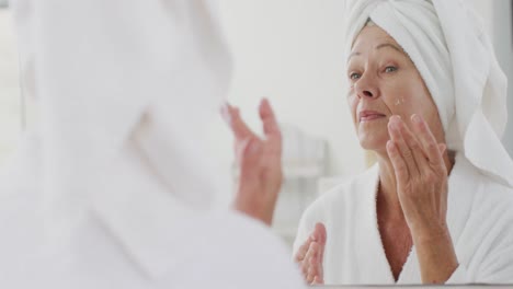 Feliz-Mujer-Caucásica-Mayor-Mirando-El-Espejo-En-El-Baño-Y-Aplicándose-Crema-En-La-Cara