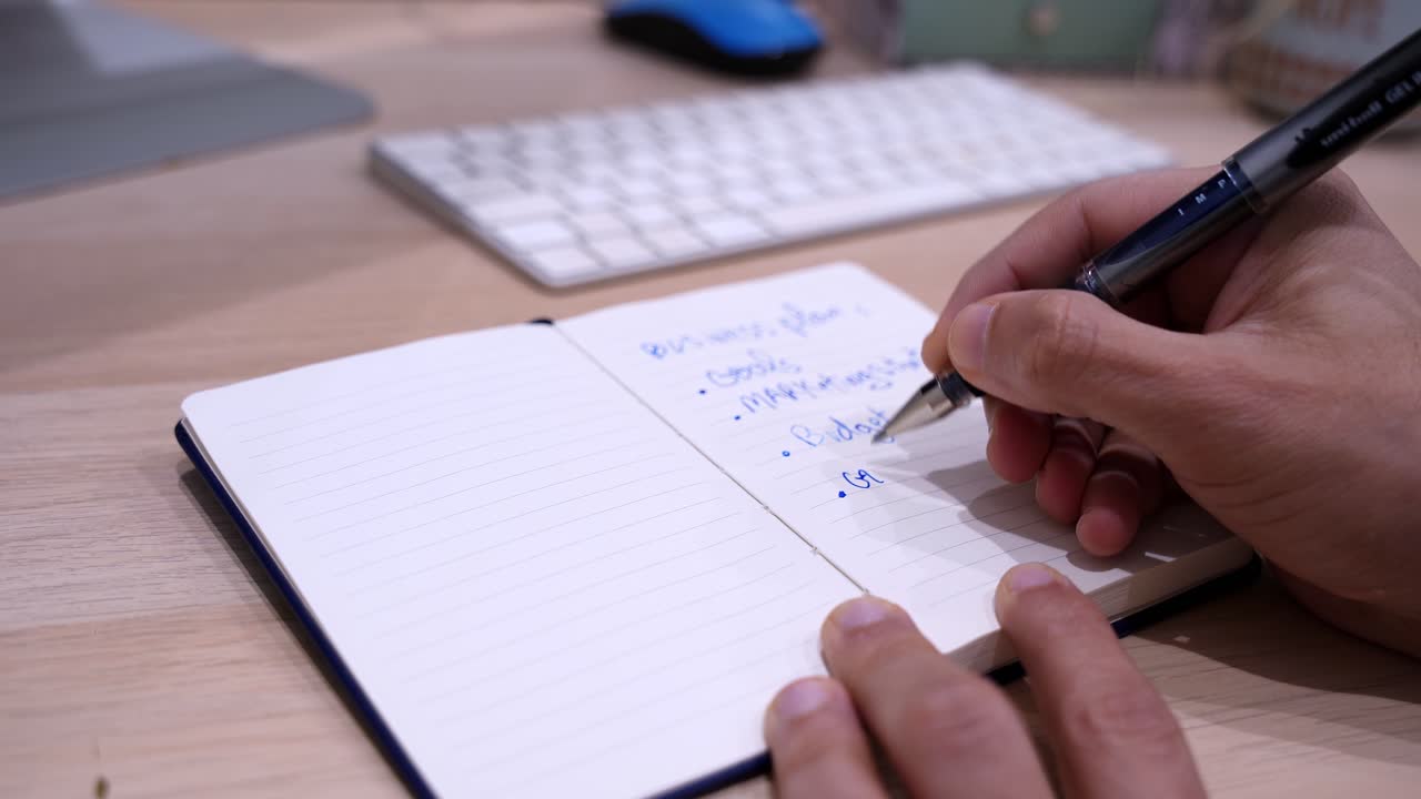 Primo piano di mani che scrivono un business plan in un taccuino aperto su una scrivania di legno, con tastiera, in luce soffusa da interni e bassa profondità di campo per concetti di startup e pianificazione