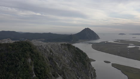 Vista-Panorámica-De-La-Montaña-Con-Cantera-Abandonada-En-Su-Cumbre-Y-Revelación-Del-Estuario-Del-Río,-La-Cresta-De-La-Montaña-En-La-Distancia-Con-Barcos-Anclados-En-La-Bahía,-Rusia