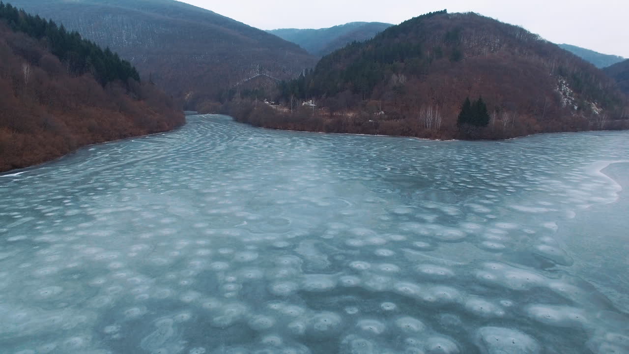 AERIAL: Drone rises above the frozen crystal ice of the lake. Lake surrounded by ok hills in winter