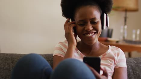 Fröhliche-Afroamerikanische-Frau-Mit-Kopfhörern-Entspannt-Sich-Auf-Der-Couch-Und-Lacht-Mit-Ihrem-Smartphone