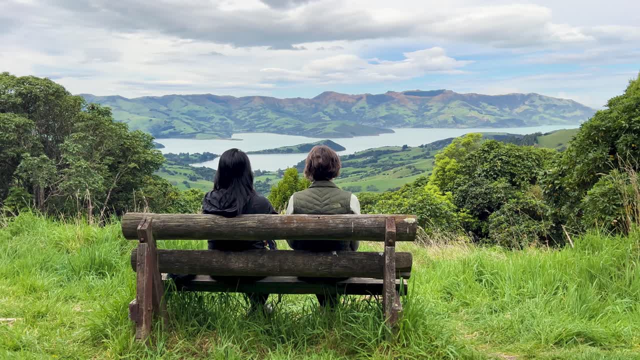 Two people enjoy scenic Akaroa Peninsula view from a bench, peaceful setting