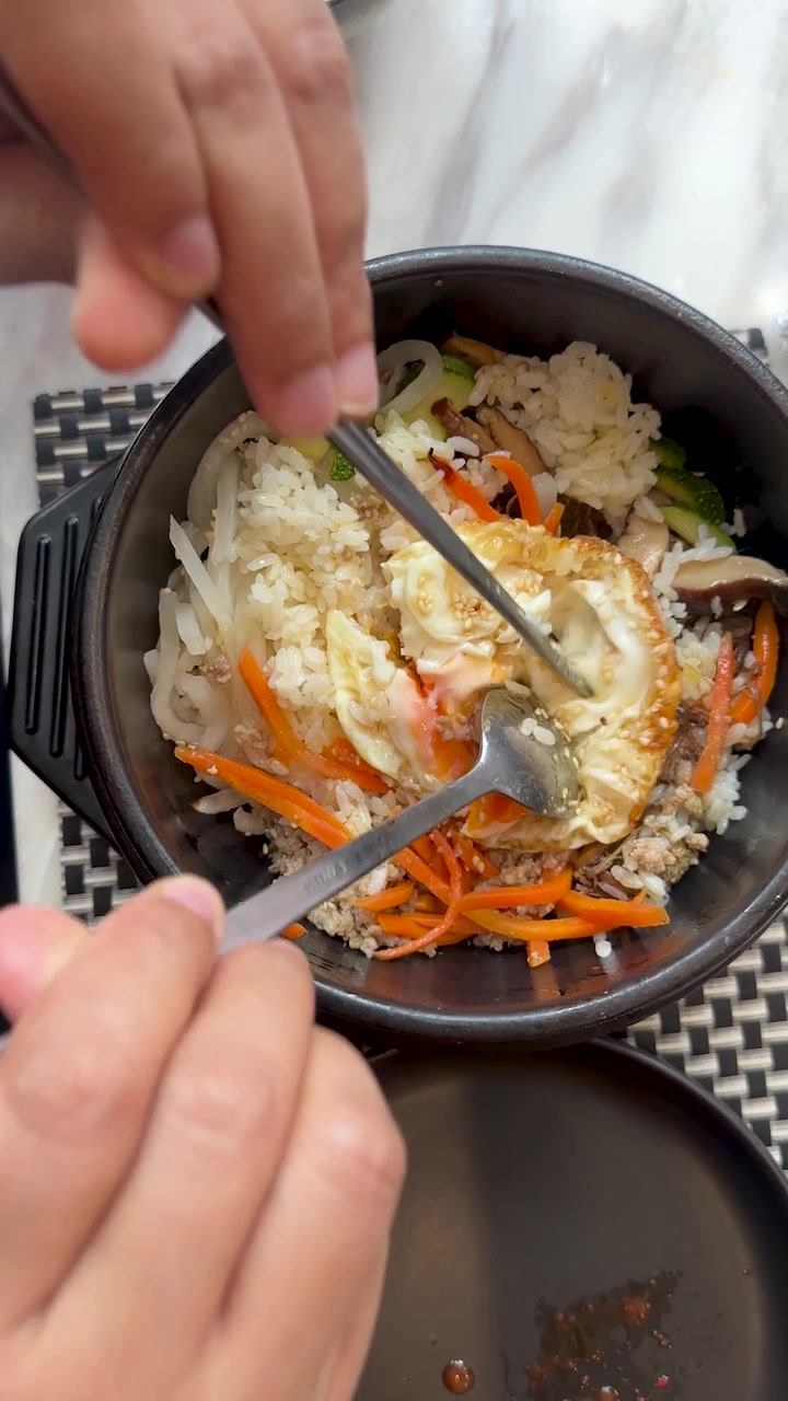 Pemandangan dari atas tangan yang menggunakan sendok dan sumpit untuk mencampur bahan bibimbap berwarna-warni