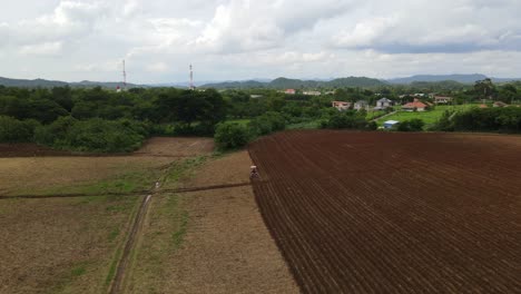 Cinematic-descending-aerial-footage-showing-a-agricultural-tractor-plowing-soil-to-prepare-for-the-next-crop-plantation-season,-a-town-and-houses-seen-mountains-visible-in-the-horizon