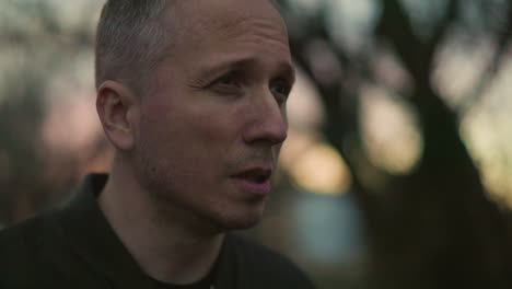 a man in a green jacket lights a cigarette outdoors at dusk, smoking, with a blur view of trees