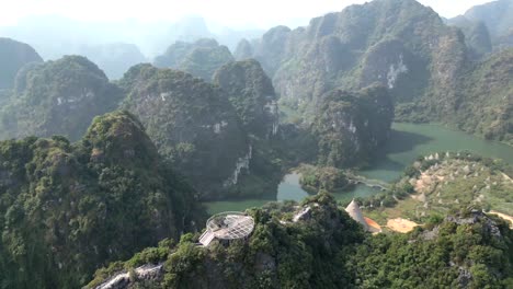 aerial video of a mountain skywalk viewpoint with glass platform, surrounded by lush cliffs, cone-shaped structures, rivers, and karst formations in the valley.