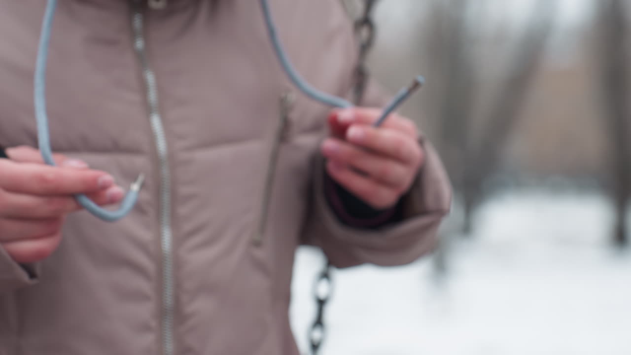 primer plano de una persona balanceándose en traje de invierno, jugando con encaje de capucha mientras agarra la cadena de balanceo, crear un entorno frío al aire libre en un parque durante el invierno