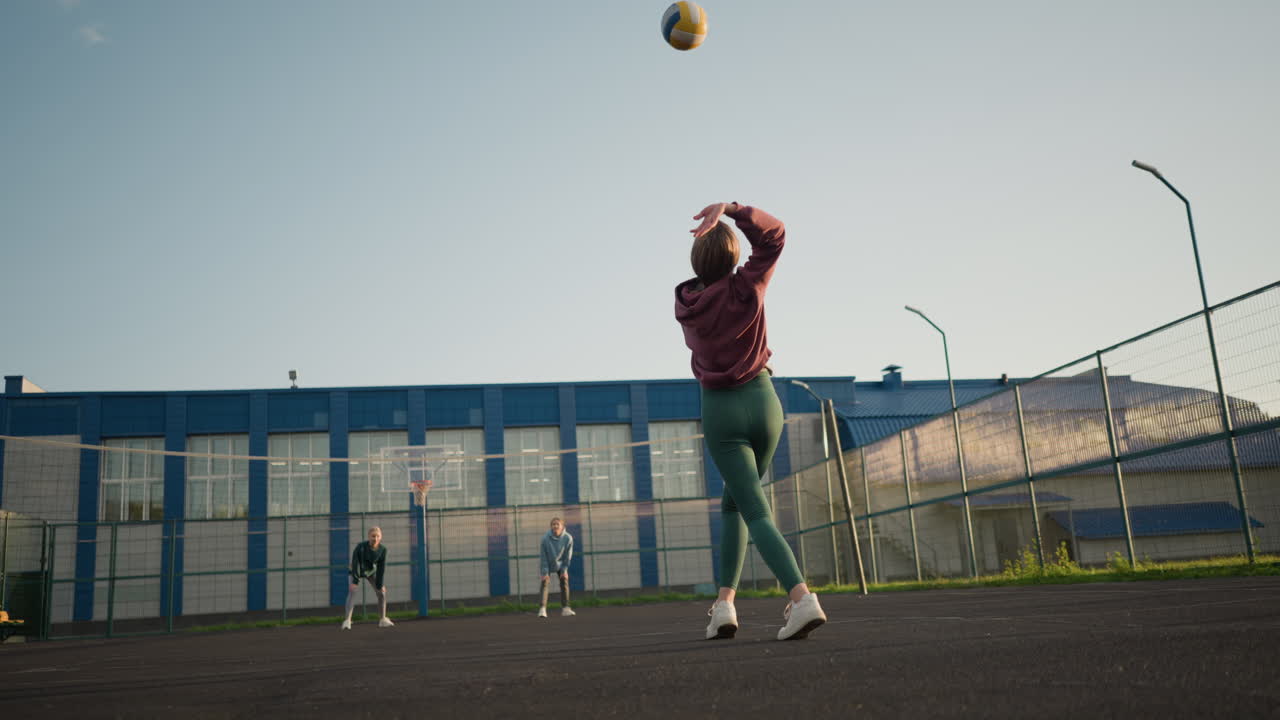 dos mujeres en el otro lado de la cancha de voleibol. la dama de leggings verdes sirve voleibol mientras la dama de capucha verde salta para atraparlo, el edificio y la cancha al aire libre visibles en el fondo