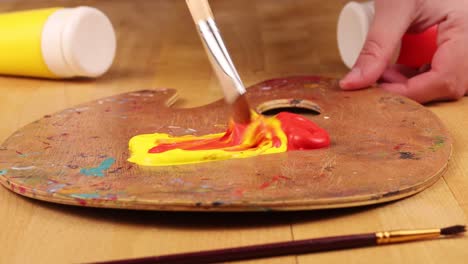 mixing paint yellow and red to create orange on a palette with paintbrush