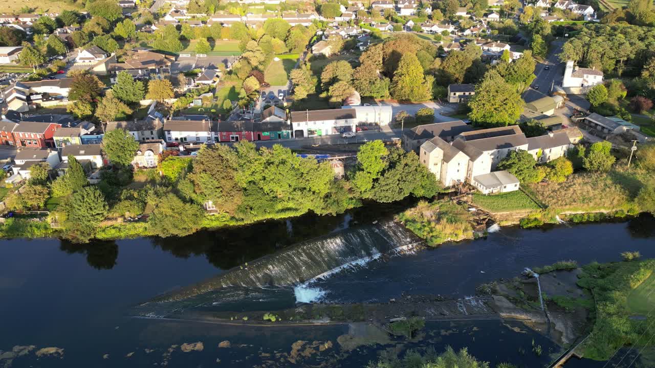 Vista aerea con drone dall'alto del fiume Nore che scorre attraverso Bennettsbridge, che mostra edifici di mulini in pietra, case del villaggio e verdi pascoli sotto la calda luce della sera