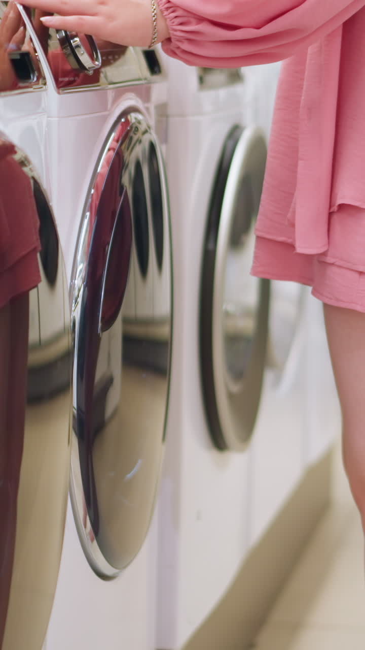 Lady in pink dress walks up to a row of washing machines in a store, closely examining them as her reflection appears on the glossy surface of the machines, with the store well lit up