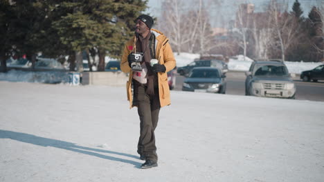 camera operator walking along snowy street holding drink cup and camera in gloved hands while looking sideways at parked cars under soft winter daylight casting long shadows over pavement