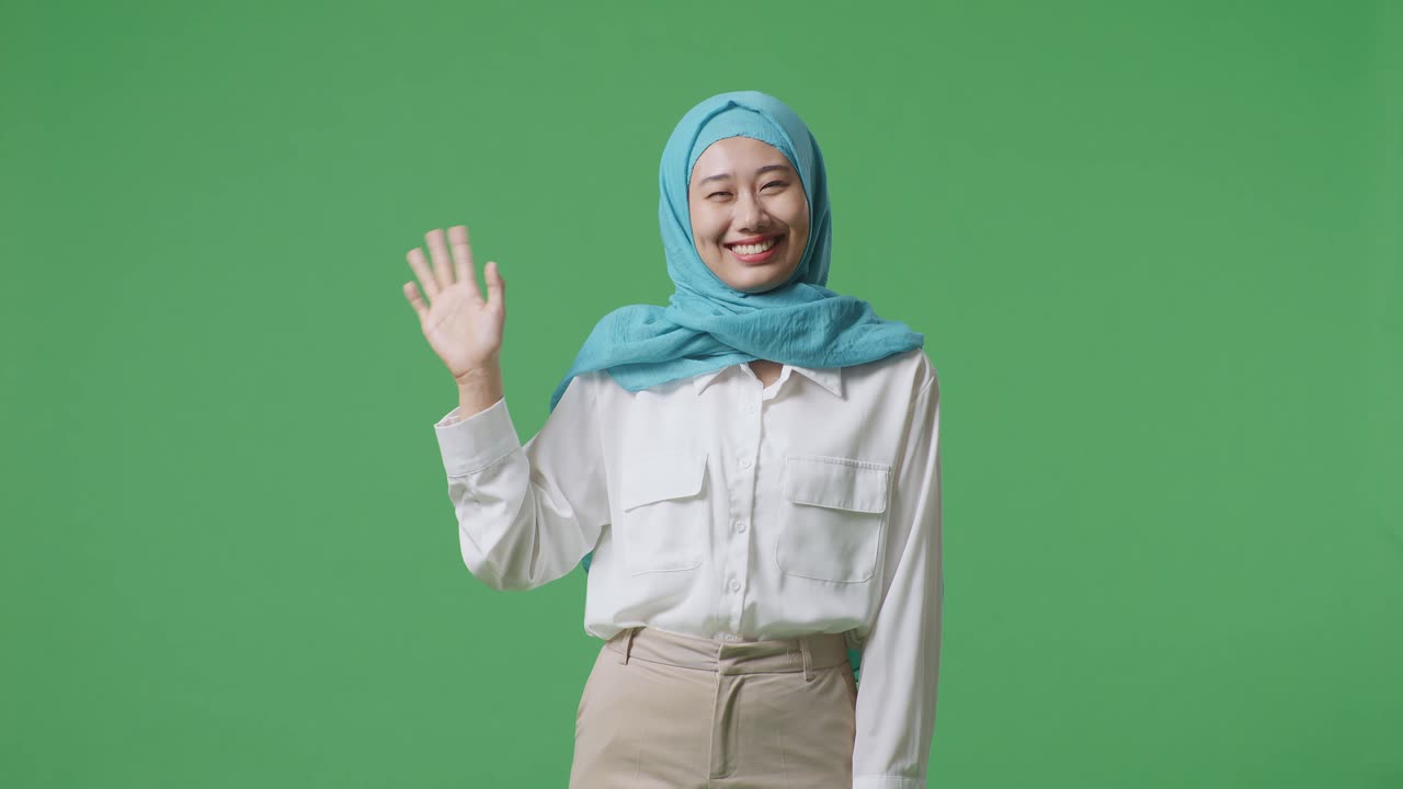 아시아의 무슬림 여성이 미소 짓고 손을 흔들며 녹색 스크린 배경 스튜디오에 서서 카메라에 인사를 하고 있습니다.