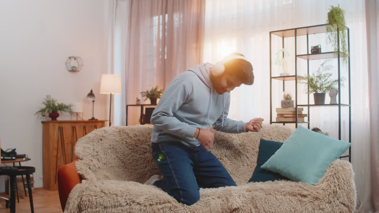 Young man dancing on couch with wireless headphones playing air guitar and enjoying disco music
