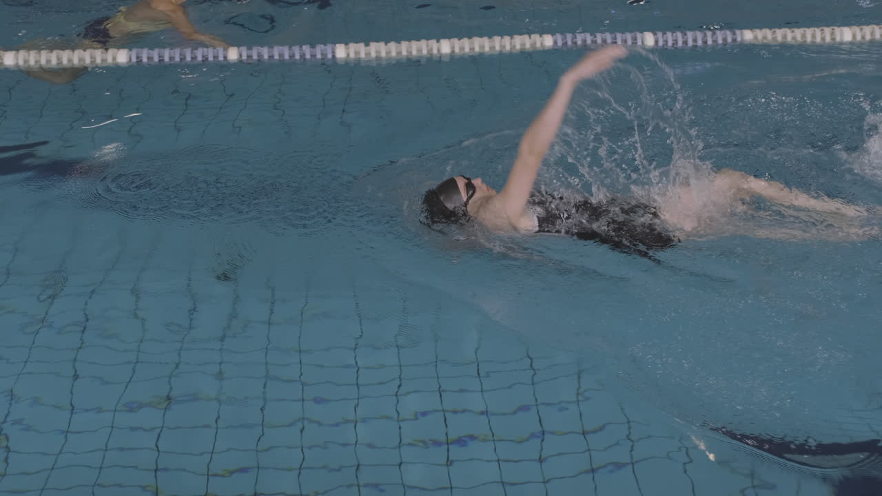 실내 수영장에서 배영을 하는 젊은 여성 수영 선수의 측면 추적 샷