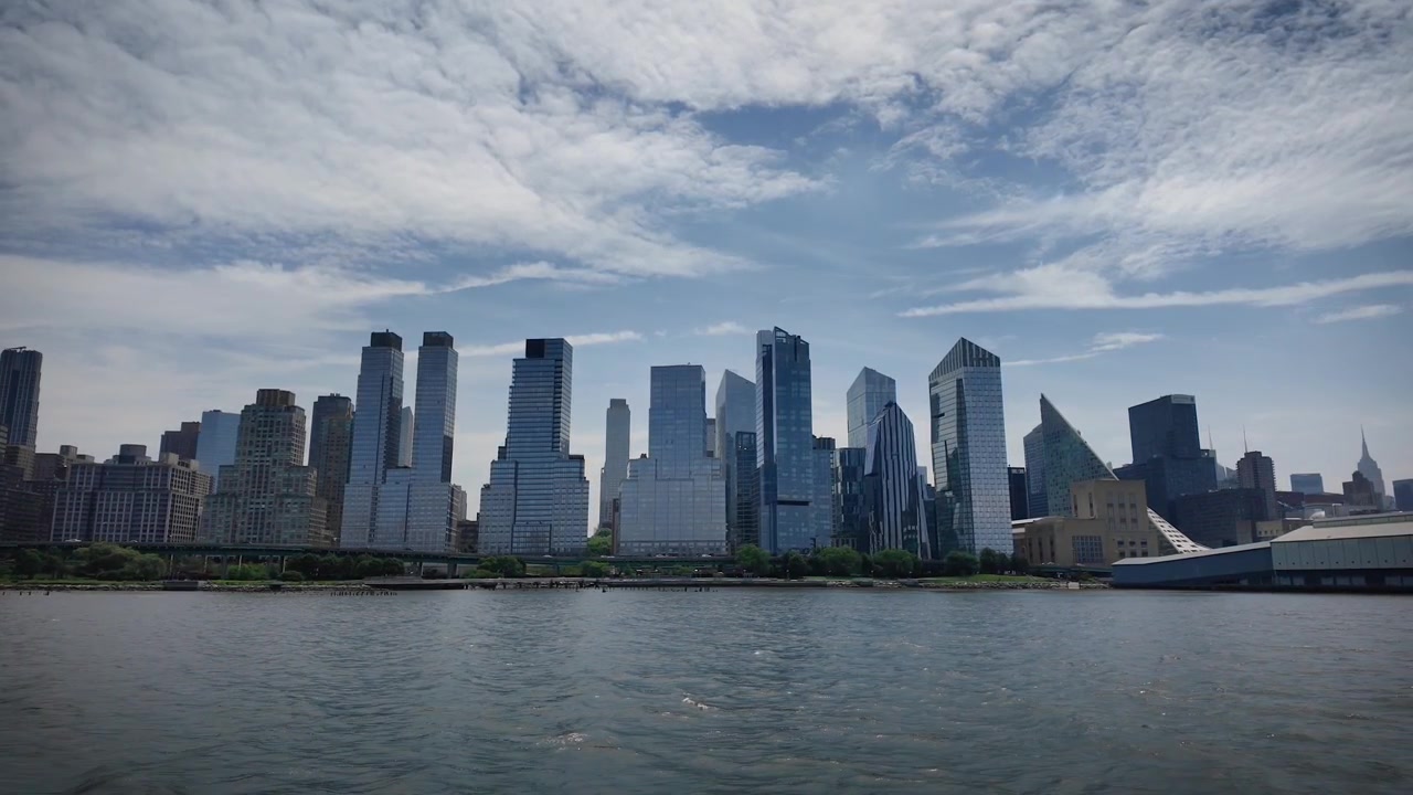 Vista cinematografica di Lower Manhattan e del One World Trade Center dal fiume Hudson, con nuvole drammatiche, acqua calma e uno skyline moderno a New York City.