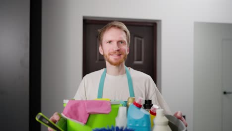 portrait of a confident blond guy in a blue apron with a large gray plastic basin with cleaning tools in a modern apartment