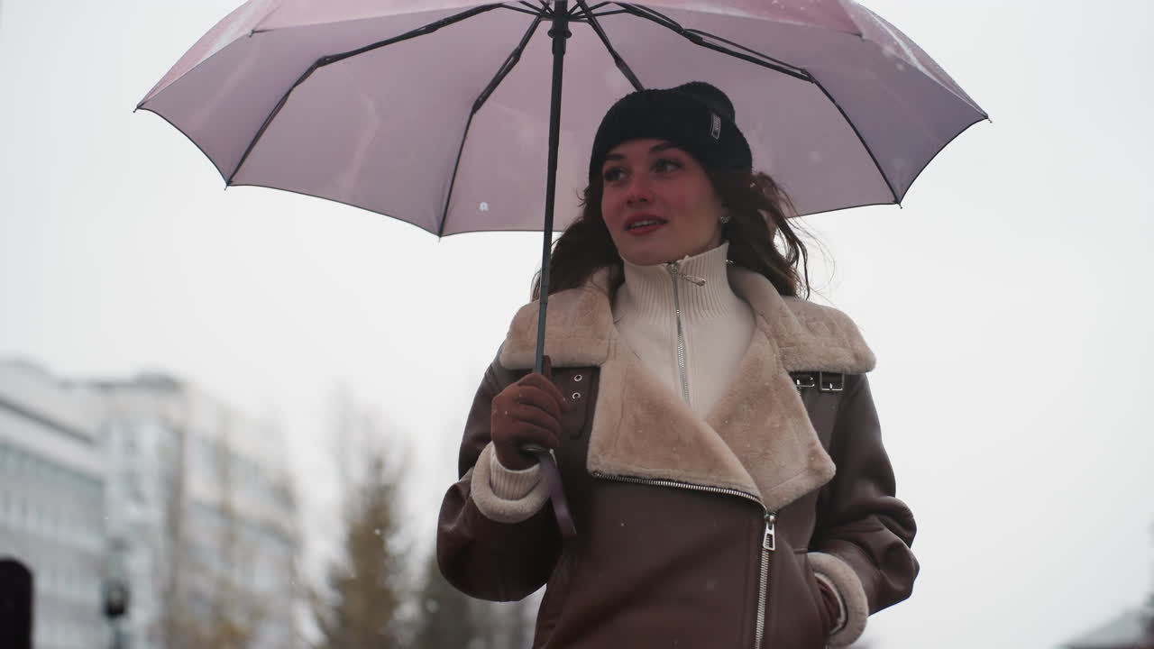 Upper body shot of lady smiling and walking in light snowfall holding umbrella wearing knit cap brown shearling jacket, cold weather, casual, outdoor, winter, city background, peaceful atmosphere