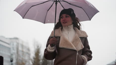 upper body shot of lady smiling and walking in light snowfall holding umbrella wearing knit cap brown shearling jacket, cold weather, casual, outdoor, winter, city background, peaceful atmosphere