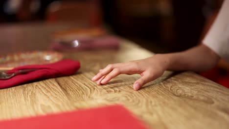 a person rhythmically taps their fingers on a wooden table beside a red napkin in a warmly lit restaurant, captured in a medium close-up shot