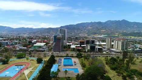 Flying-over-the-La-Sabana-park,-inside-the-city-on-San-José-in-Costa-Rica,-looking-at-sports-fields-and-tall-buildings