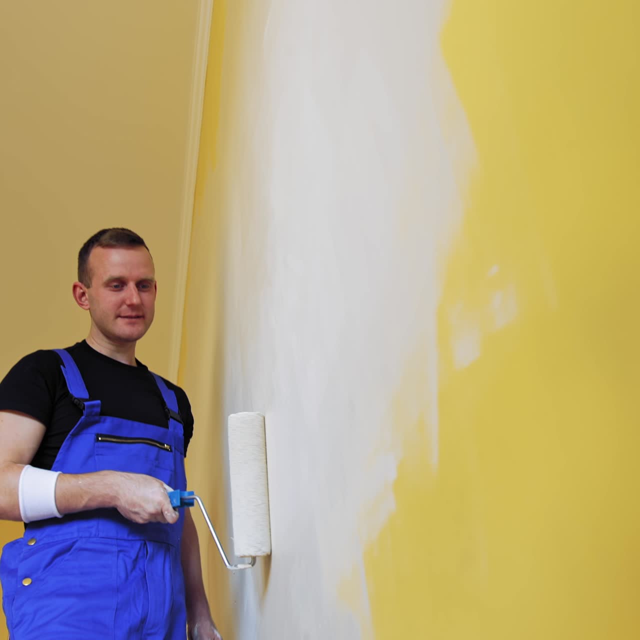 Roller repainting wall into light color. Young man in blue overalls painting wall into white in the apartment. Makeover in flat