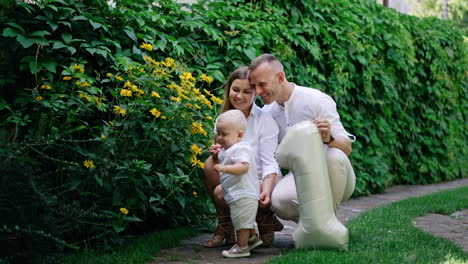 cute little baby boy stands at the yellow flowers growing outdoors. mom and dad sit squatted beside holding a balloon of number one shape.