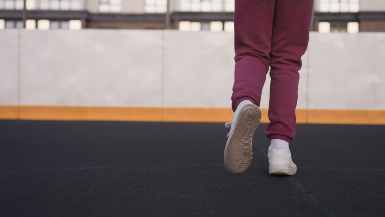 Leg view of wellness advocate jogging round barbed wire sports court with maroon tracksuit trousers and white running shoes on black asphalt surface concrete wall topped with fence urban background