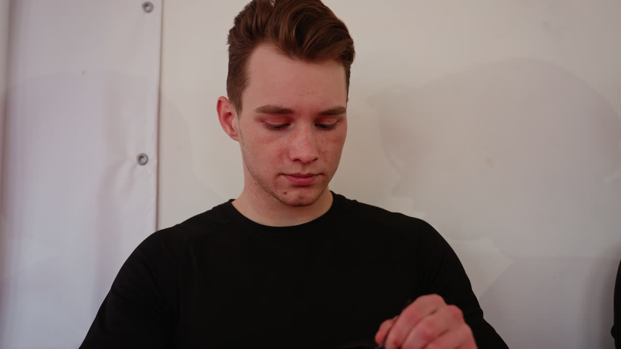 Young man in black uniform with focused downward gaze showing expression of thought, concentration, and seriousness indoors against plain background, in training atmosphere