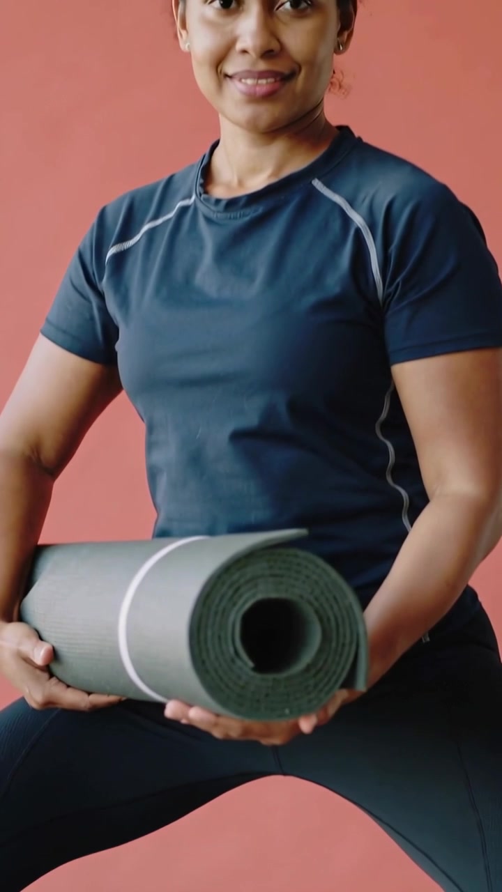 Video vertical: Mujer en leggings sosteniendo una colchoneta enrollada, la cámara asciende en un escenario coral, preparándose.