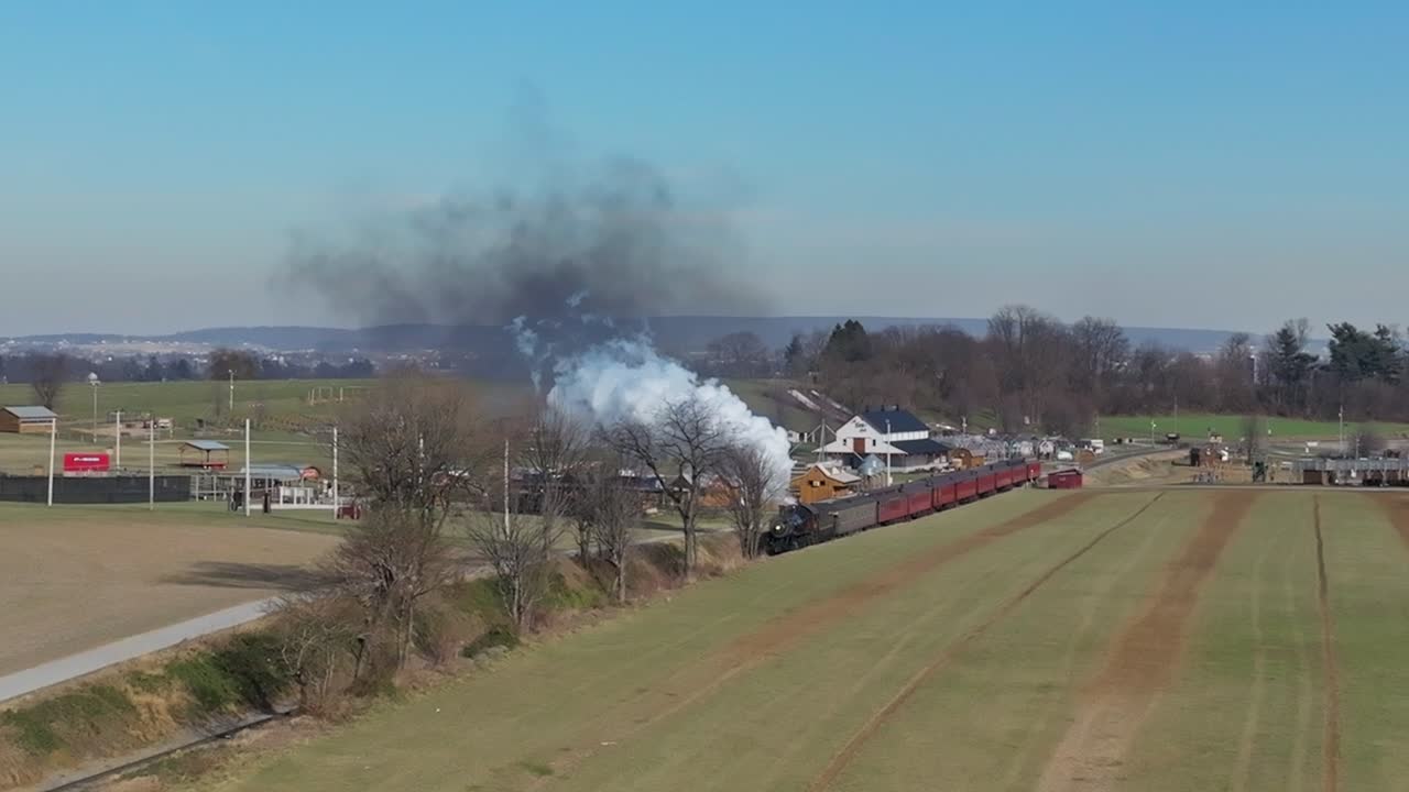 una vista aérea de un tren de pasajeros de vapor que se acerca, en cámara lenta, soplando humo, mientras viaja por el campo, en un soleado día de invierno