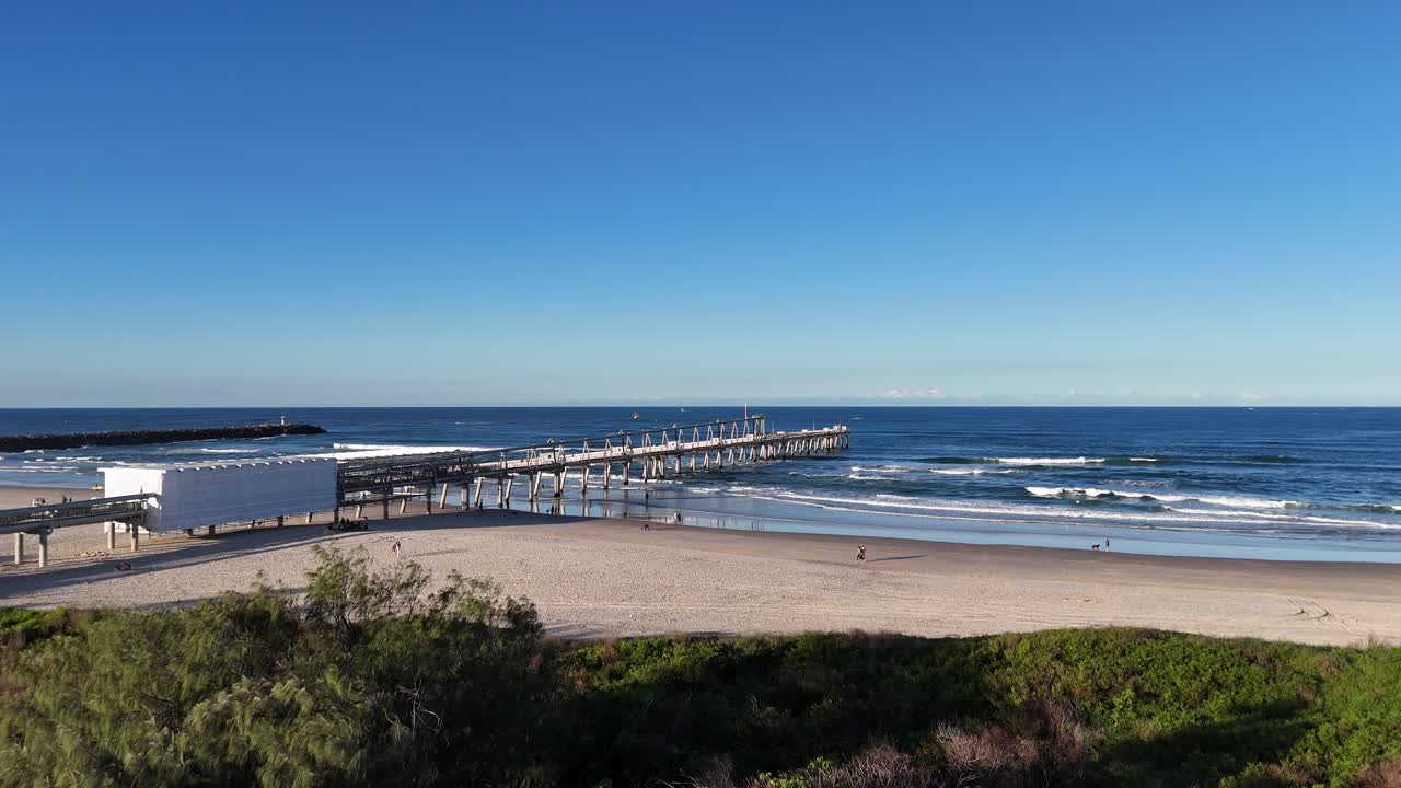 Drone footage captures a serene Gold Coast beach and jetty under clear skies, highlighting the expansive ocean and coastal landscape