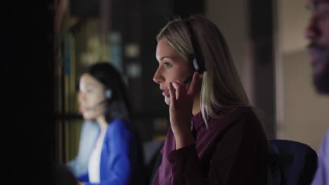video of smiling caucasian businesswoman and colleagues using headsets at night working in office