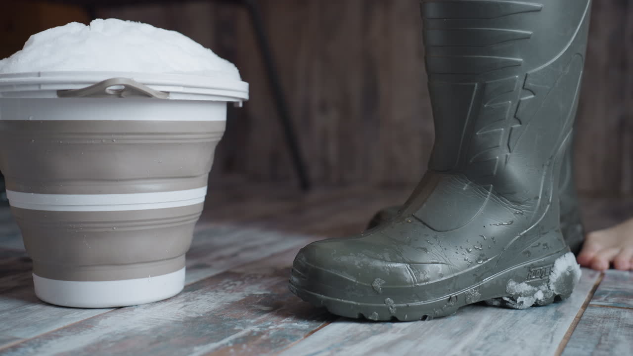 Owner removing boots touched with snow as she places them by corner of wooden entryway, melting droplets pooling on rustic tiled floor, soft winter light casting gentle shadows across threshold