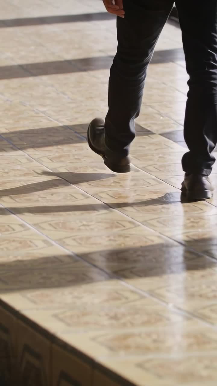 Video verticale: Uomo in pantaloni scuri che scende da una sporgenza camminando su piastrelle verso una porta, con fasce d'ombra.
