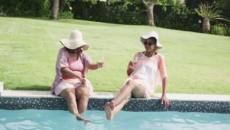 happy senior african american female friends in sunhats sitting by pool with drinks, slow motion