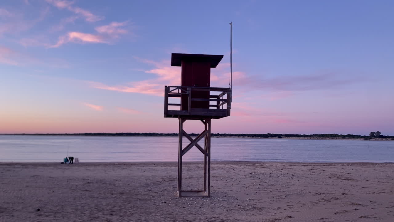 Livreddertårn våger over en rolig sandstrand og stille vand under en farverig solnedgang i Sanlucar de Barrameda