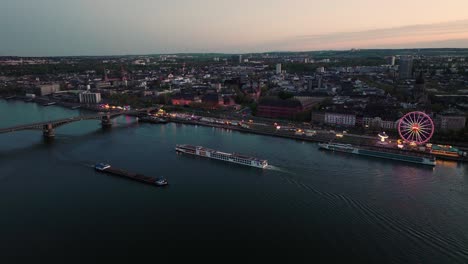toma aérea de drones de mainz dos barcos cruzando frente a un festival de vino de verano en la orilla del río rin bajo el cielo naranja de la puesta de sol