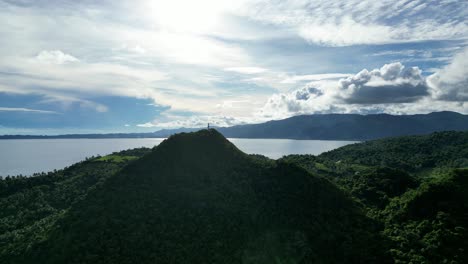 a high contrast aerial view of the bote lighthouse on a tree-covered hill in catanduanes, philippines, overlooking the ocean and the blue sky, with dramatic clouds and backlit by the sun