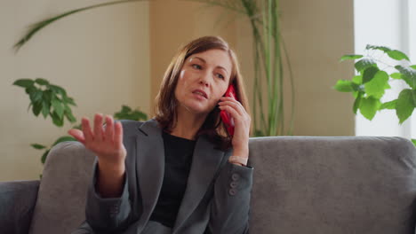 frustrated businesswoman in gray suit talking on phone indoors gesturing emotionally while sitting on couch near green plants in modern office or home environment during daytime
