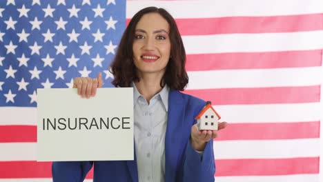 woman holding insurance sign and model house in front of us flag
