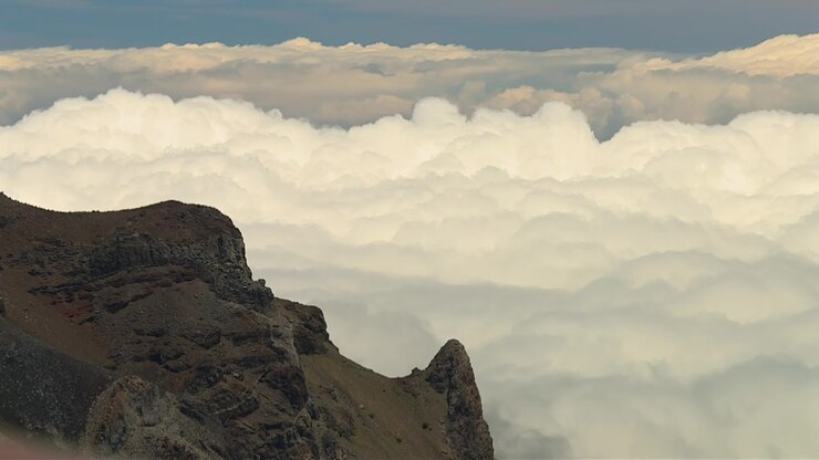 Filmato statico mozzafiato di aspre scogliere vulcaniche nel Parco Nazionale di Haleakala che si ergono sopra un vasto mare di nuvole sotto una luce diurna soffusa, mostrando un terreno drammatico e ampie vedute del cielo.