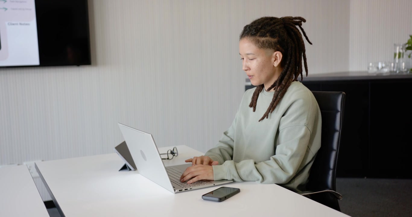 Nicht-binäre afroamerikanische Person tippt auf Laptop an langem weißen Besprechungstisch, blickt auf Folie, mit Freiraum.