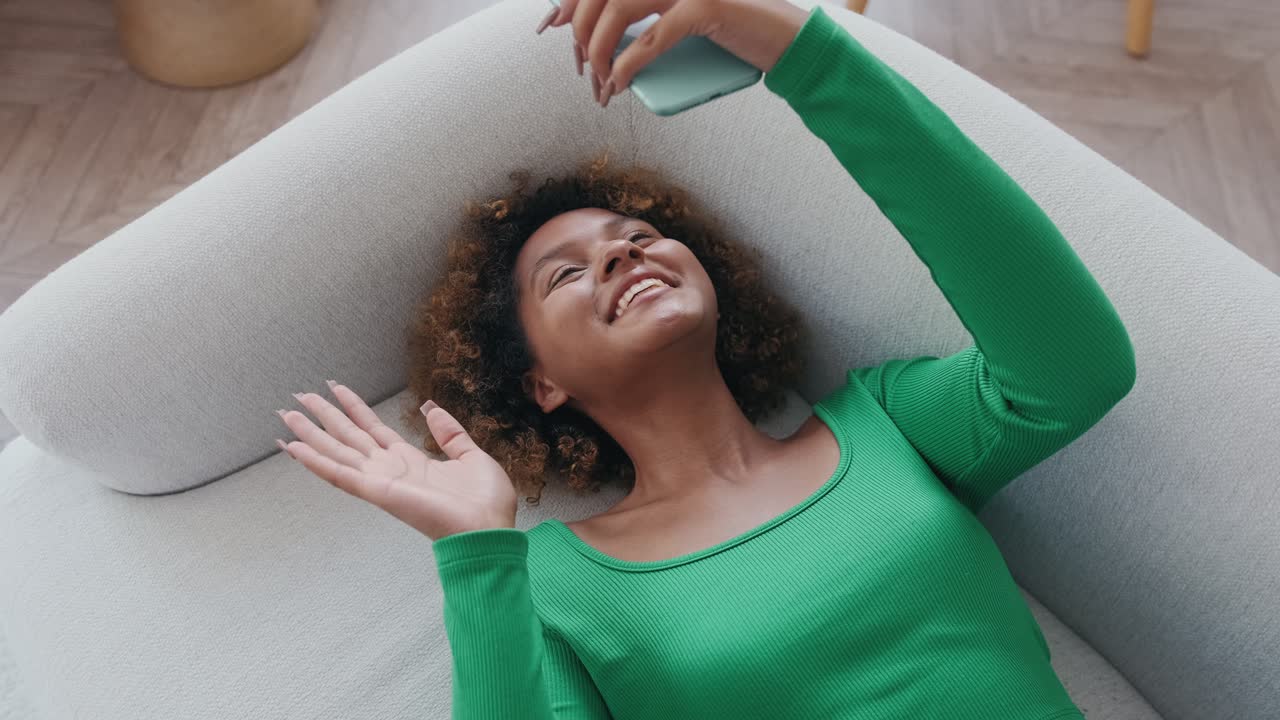una joven afroamericana alegre hace una llamada de video en el teléfono y se acuesta en el sofá.