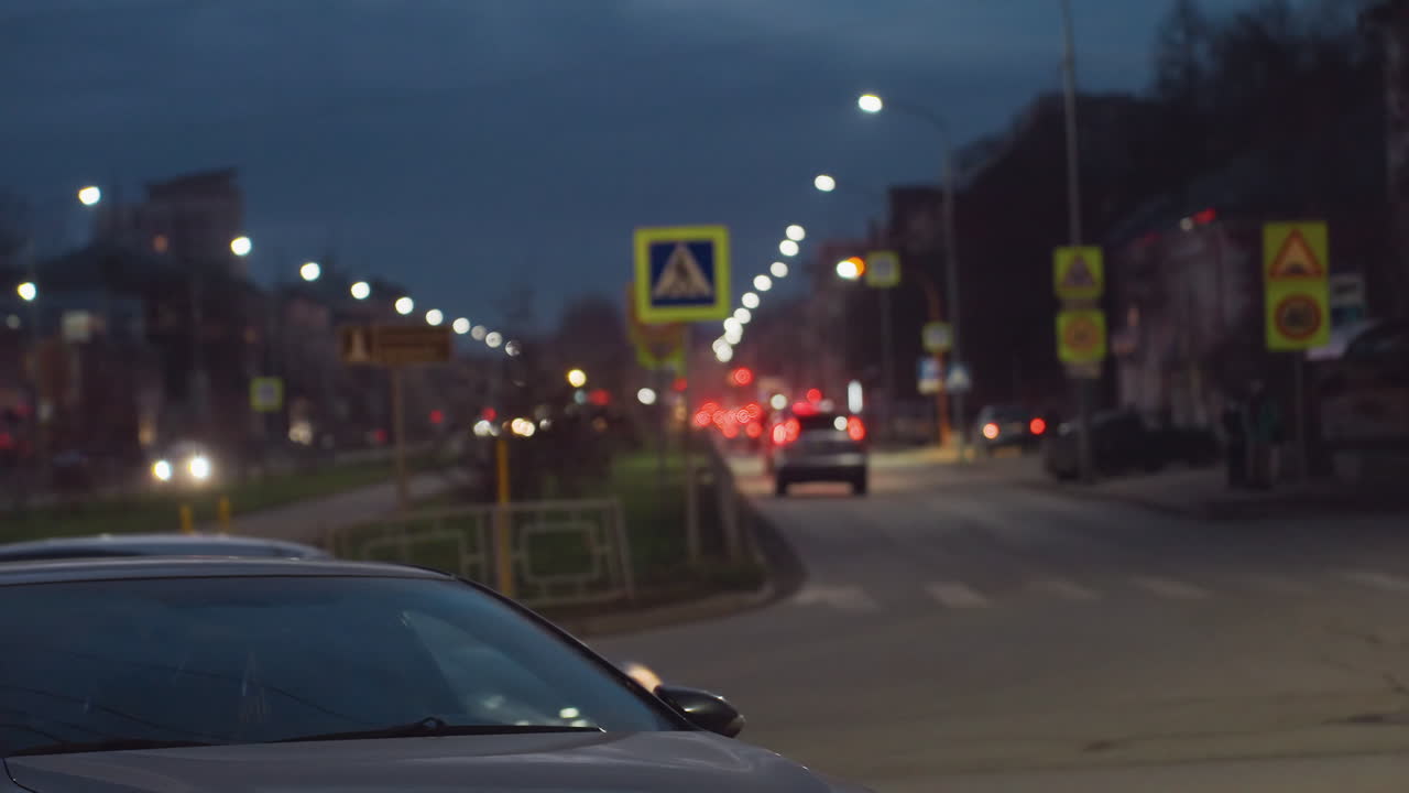 Busy night road scene with headlights of passing cars creating light trail along street lined with road signs and lampposts, parked car visible at corner under dark evening sky