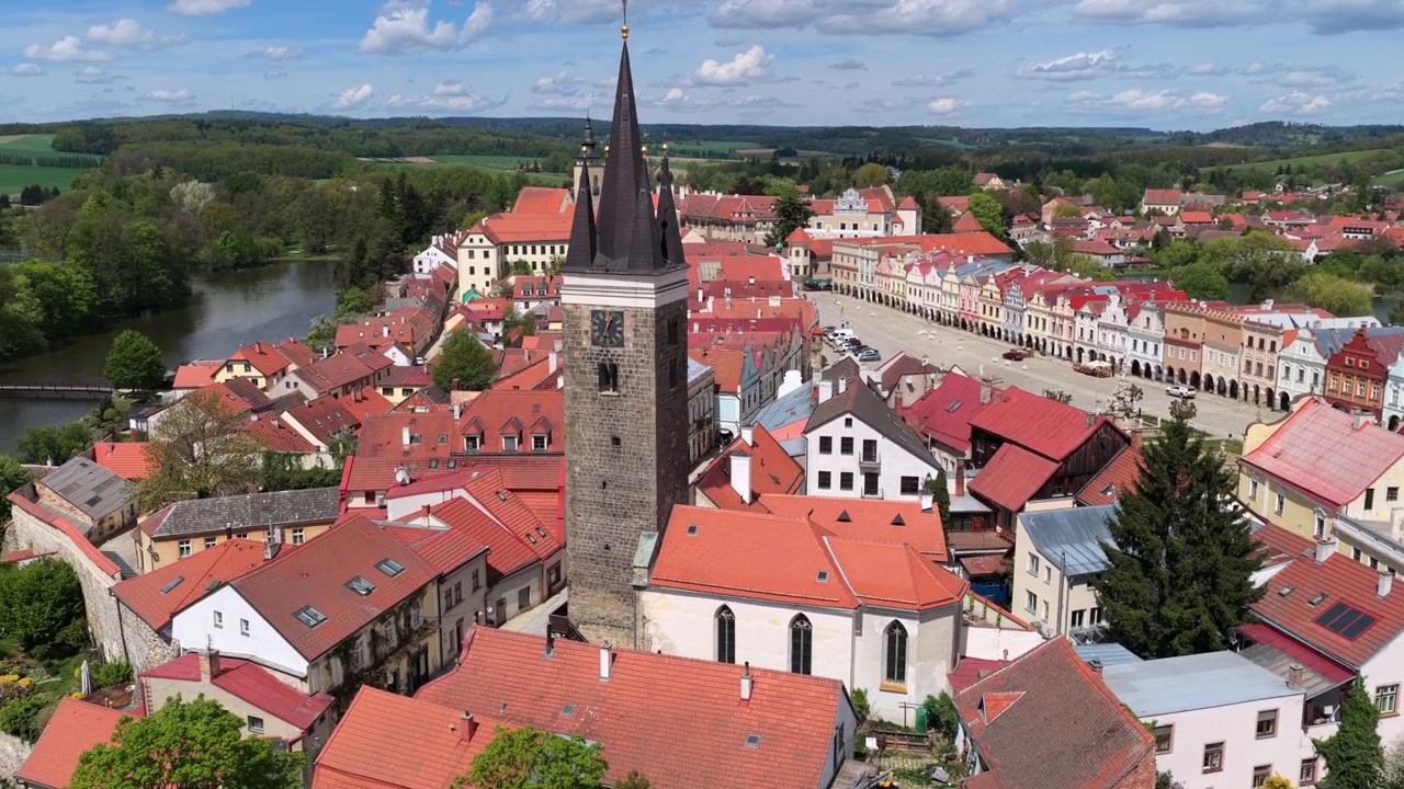 Vista aérea de drone de uma alta torre do relógio gótica elevando-se acima de uma praça histórica tcheca com casas coloridas com arcadas, torres de igreja e paisagem rural ondulante ao fundo, Telč, República Tcheca