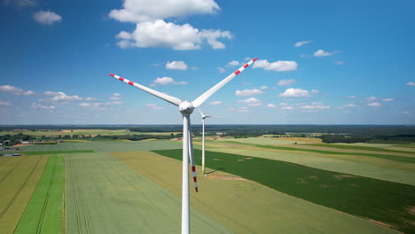 Slowly-rotating-wind-turbine-blades-in-a-beautiful-sunny-setting-against-a-lightly-cloudy-blue-sky-and-ripening-green-fields