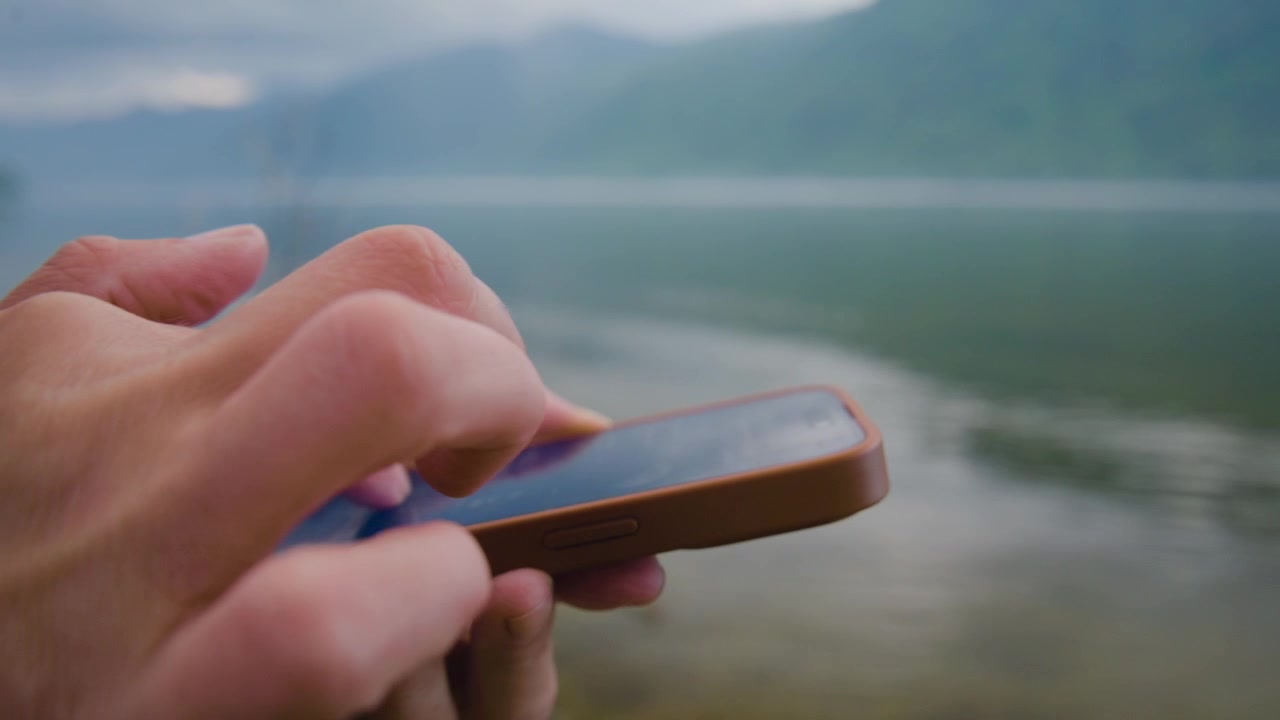 Mani scorrono il telefono vicino a un lago calmo. Le dita toccano lo schermo con concentrazione. Montagne nebbiose incombono dietro l'acqua. Un momento tranquillo all'aperto, connessione digitale. Ottimo per temi di viaggio, natura, tecnologia o consapevolezza.