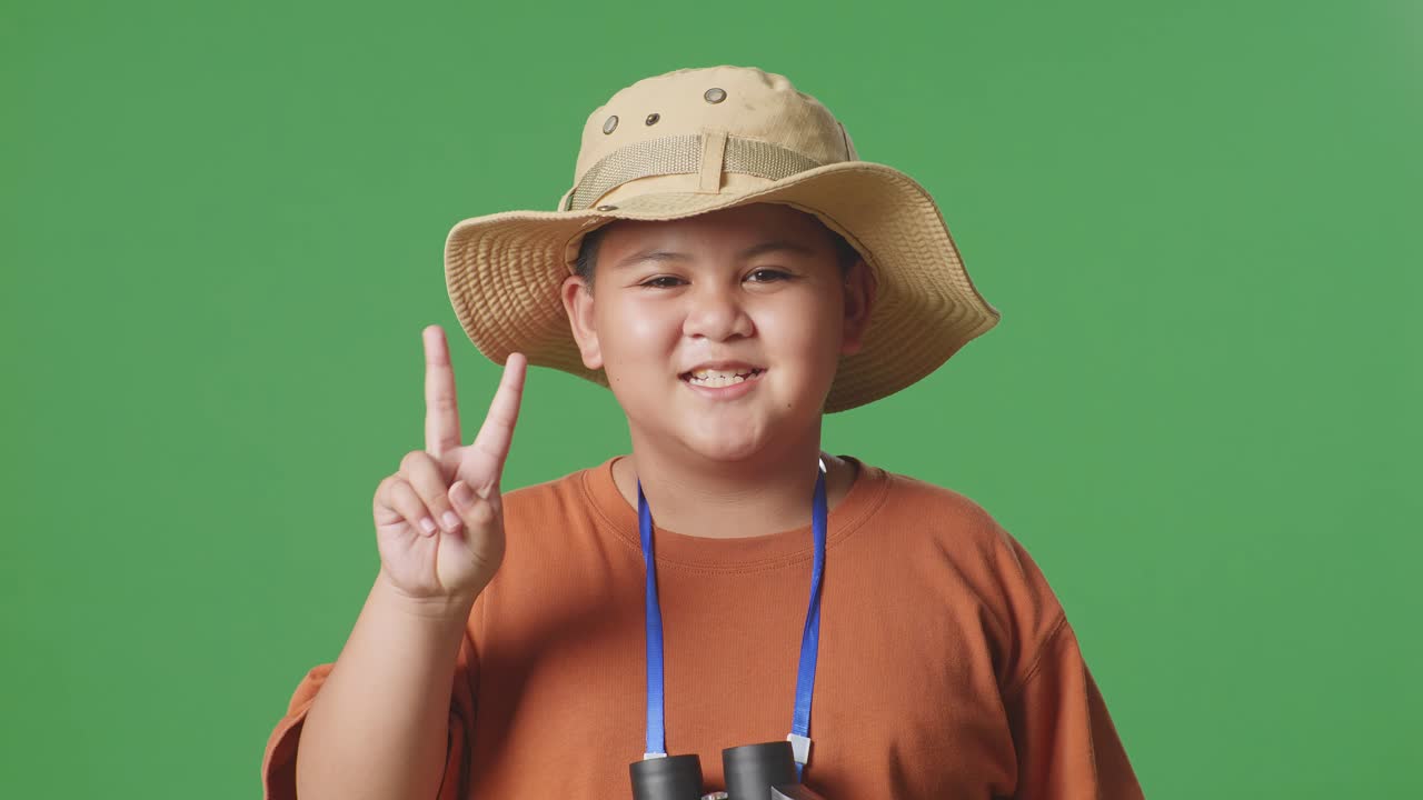 모자와 안경을 쓴 아시아 관광 소년은 보기를 사용하고 녹색 스크린 배경에서 평화의 몸짓을 보여줍니다. 소년 연구자는 무언가를 조사합니다.